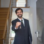 Smiling realtor in a black suit handing keys through an open door inside a modern house.