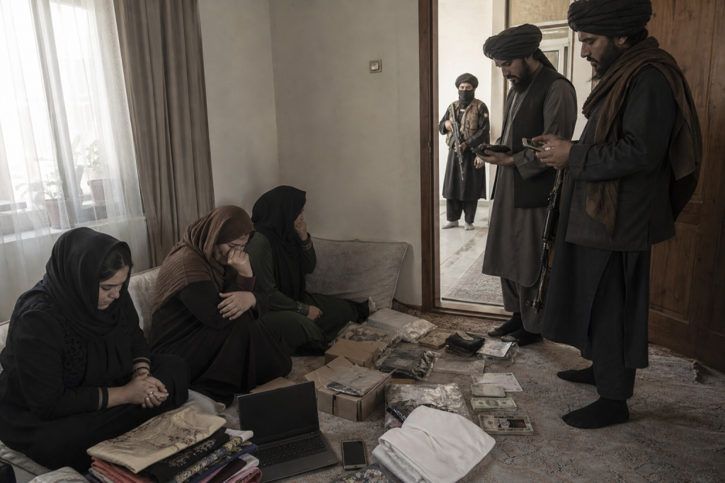 Three women sit on a floor amid clothing, electronics, documents, and cash while three armed men stand nearby