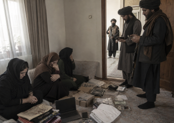 Three women sit on a floor amid clothing, electronics, documents, and cash while three armed men stand nearby