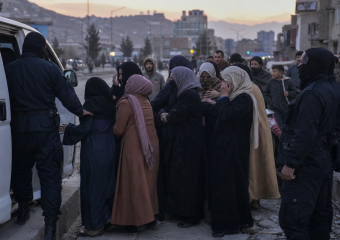 Women in head coverings gather beside a white van as uniformed officers assist them in an urban street