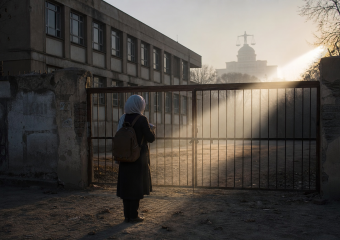 Person wearing a headscarf and backpack stands before a closed gate, with a courthouse visible in the background